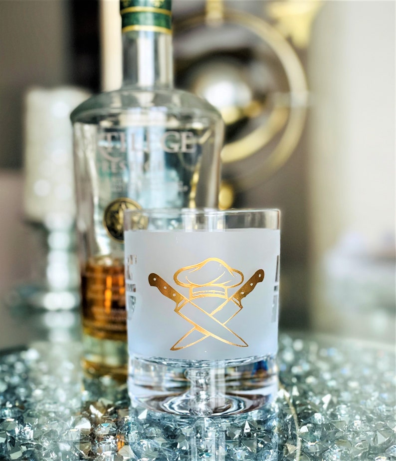 May include: A frosted glass with a gold chef hat and crossed knives design. The glass is sitting on a table with a bottle of liquor in the background.