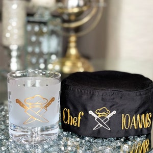 May include: A black chef's hat with gold embroidery that says "Chef Ioannis" and a crossed chef's knife and hat design. A frosted glass with a gold chef's hat and crossed knives design is in the foreground.