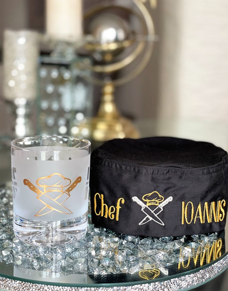 May include: A black chef's hat with gold embroidery that says "Chef Ioannis" and a crossed chef's knife and hat design. A frosted glass with a gold chef's hat and crossed knives design is in the foreground.