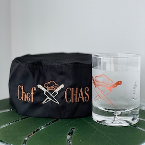 May include: Black chef hat with the text "Chef Chas" embroidered in brown thread. The hat has a white chef hat and crossed knives design. A frosted glass with a similar design is next to the hat.
