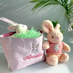 May include: Pink and white striped Easter basket with a green grass filler. The basket has the name "Annelise" embroidered on the front. A white bunny and a pink bunny are in the basket.
