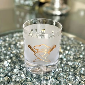 May include: A frosted glass with a gold chef hat and crossed knives design. The glass is sitting on a bed of sparkly gold stones.