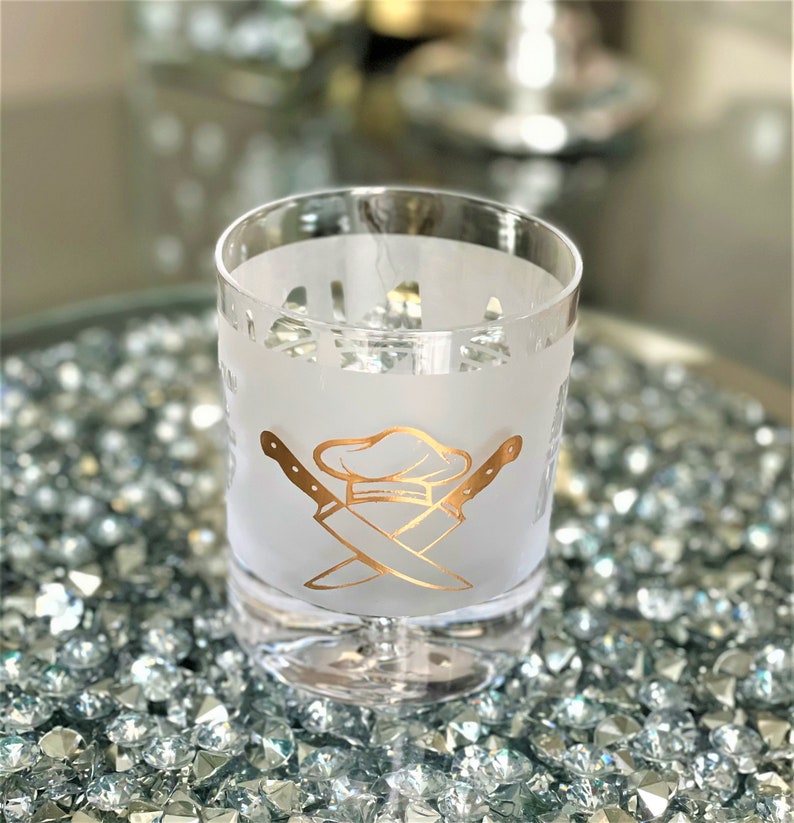 May include: A frosted glass with a gold chef hat and crossed knives design. The glass is sitting on a bed of sparkly gold stones.