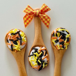 May include: Three wooden spoons with white chocolate and orange, black, and yellow sprinkles. The spoons are arranged in a triangle shape with a yellow and orange checkered bow on top.