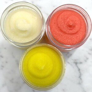 Puede incluir: Tres frascos de vidrio llenos de exfoliantes de az&uacute;car de diferentes colores. Los frascos est&aacute;n sobre una superficie de m&aacute;rmol blanco. Los exfoliantes son amarillos, blancos y rosados.