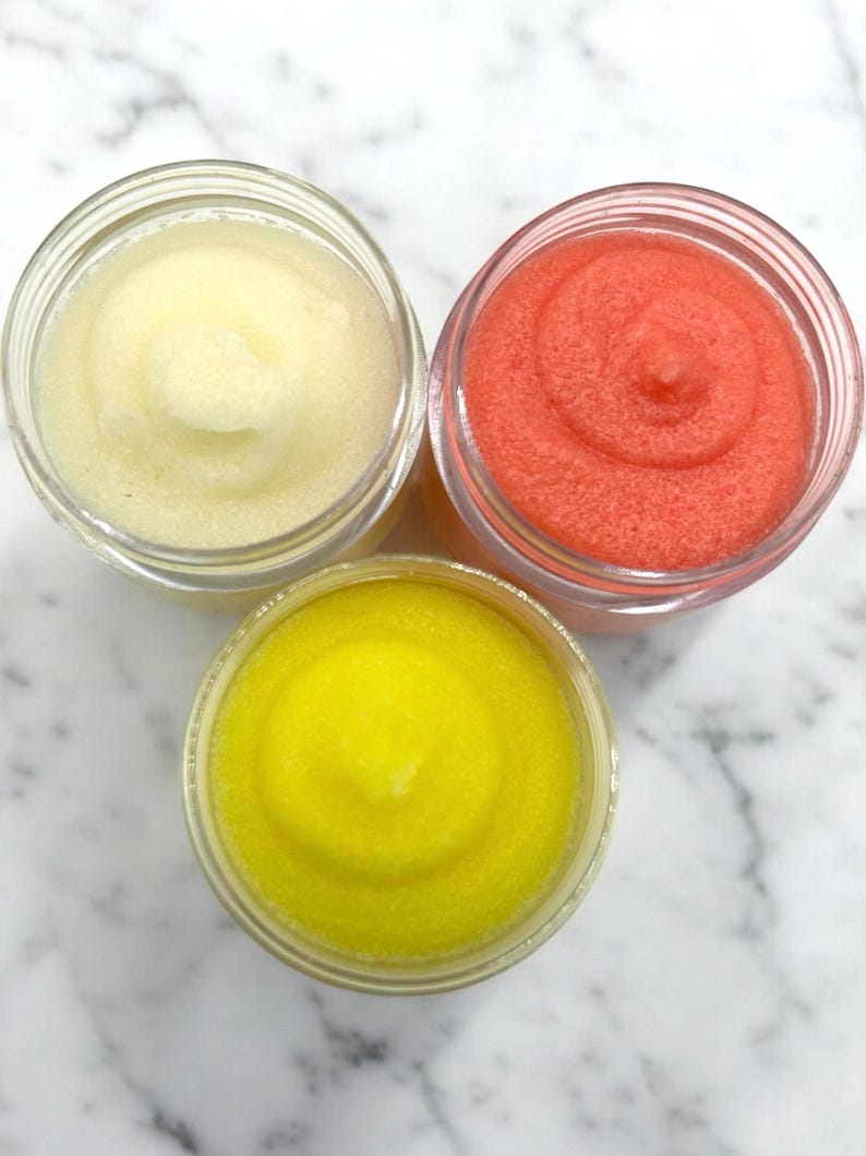 Puede incluir: Tres frascos de vidrio llenos de exfoliantes de az&uacute;car de diferentes colores. Los frascos est&aacute;n sobre una superficie de m&aacute;rmol blanco. Los exfoliantes son amarillos, blancos y rosados.