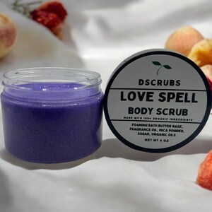 May include: A purple body scrub in a clear jar with a black lid. The jar is labeled "LOVE SPELL BODY SCRUB" with the text "MADE WITH 100% ORGANIC INGREDIENTS" and "NET WT 4 OZ".