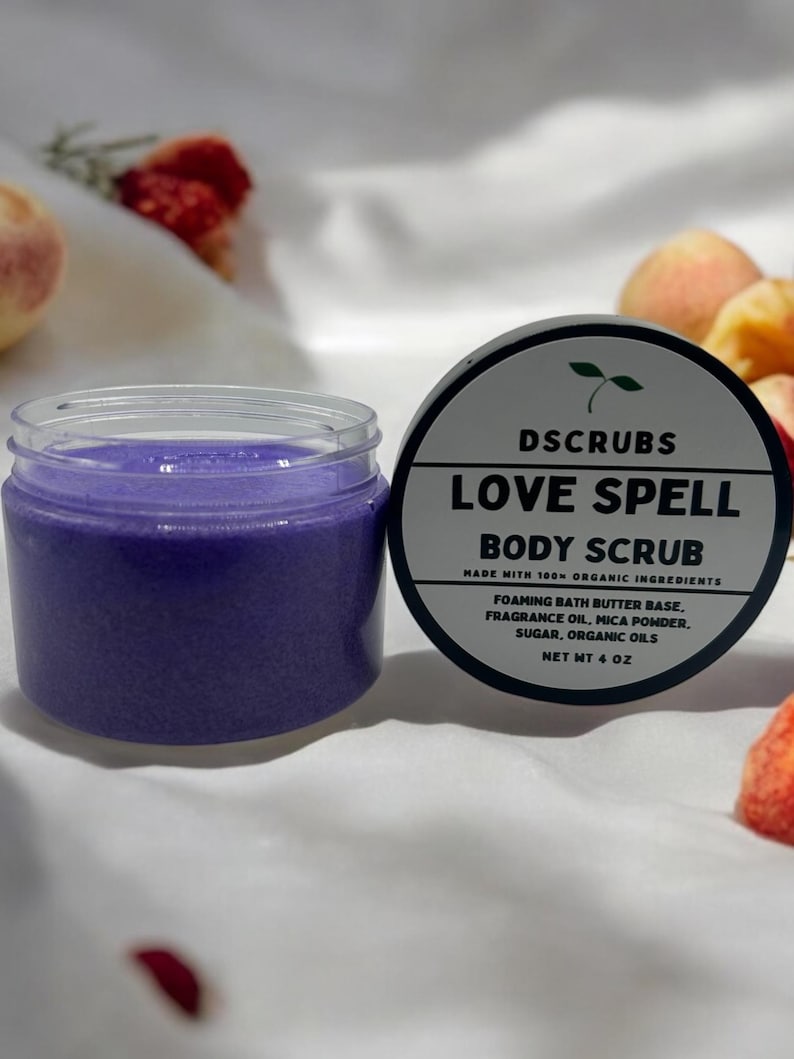 May include: A purple body scrub in a clear jar with a black lid. The jar is labeled "LOVE SPELL BODY SCRUB" with the text "MADE WITH 100% ORGANIC INGREDIENTS" and "NET WT 4 OZ".