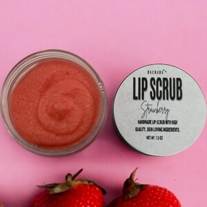 Puede incluir: Un peque&ntilde;o tarro de exfoliante labial rosa con el texto "DSCRUBS LIP SCRUB Strawberry HANDMADE LIP SCRUB WITH HIGH QUALITY, SKIN LOVING INGREDIENTS. NET WT. 1.3 OZ" en la etiqueta. El tarro est&aacute; sobre una superficie rosa con fresas alrededor.