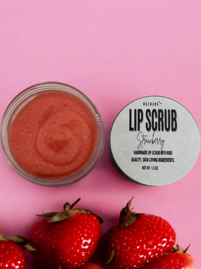 Puede incluir: Un peque&ntilde;o tarro de exfoliante labial rosa con el texto "DSCRUBS LIP SCRUB Strawberry HANDMADE LIP SCRUB WITH HIGH QUALITY, SKIN LOVING INGREDIENTS. NET WT. 1.3 OZ" en la etiqueta. El tarro est&aacute; sobre una superficie rosa con fresas alrededor.