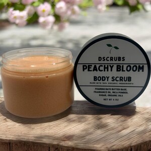 Op de afbeelding: Peachy Bloom body scrub in een glazen pot met een perzikkleurige scrub. De pot staat op een houten oppervlak. Het etiket op de pot luidt "DSCRUBS PEACHY BLOOM BODY SCRUB MADE WITH 100% ORGANIC INGREDIENTS FOAMING BATH BUTTER BASE, FRAGRANCE OIL, MICA POWDER, SUGAR, ORGANIC OILS NET WT 4 OZ"