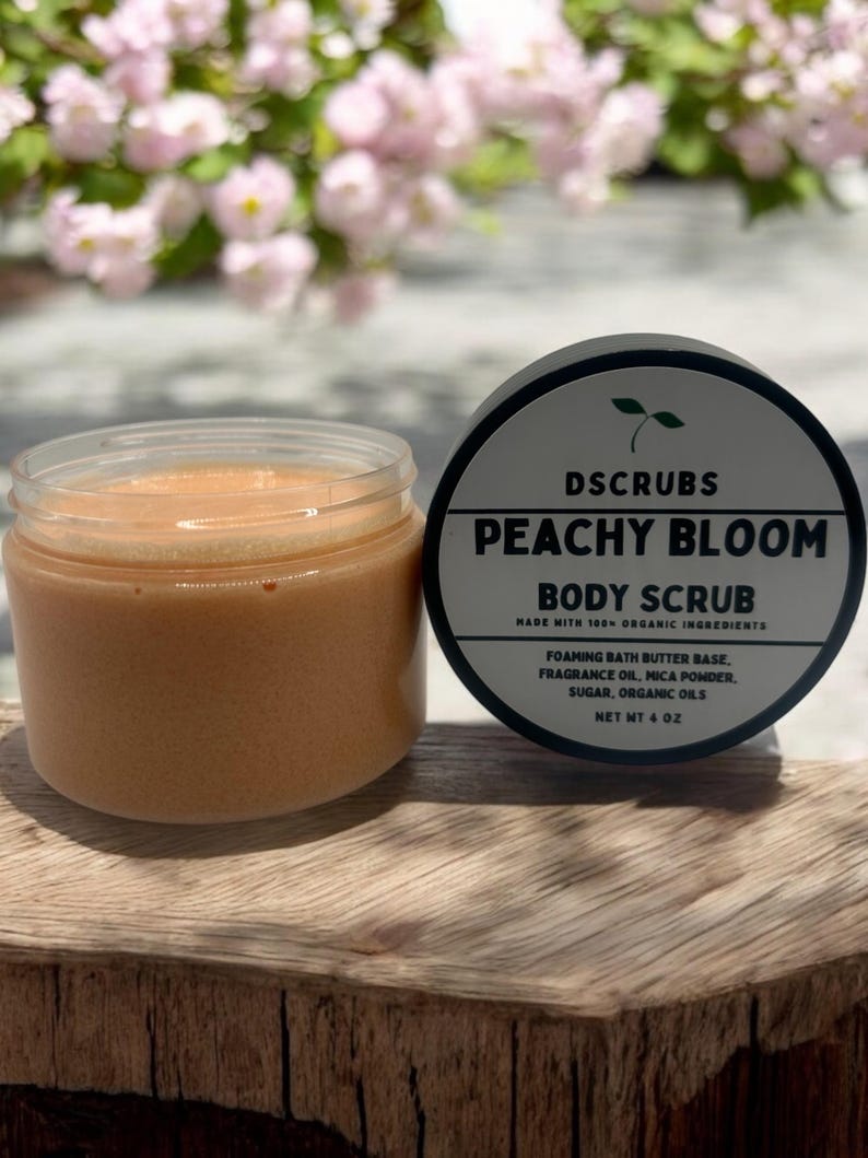 Op de afbeelding: Peachy Bloom body scrub in een glazen pot met een perzikkleurige scrub. De pot staat op een houten oppervlak. Het etiket op de pot luidt "DSCRUBS PEACHY BLOOM BODY SCRUB MADE WITH 100% ORGANIC INGREDIENTS FOAMING BATH BUTTER BASE, FRAGRANCE OIL, MICA POWDER, SUGAR, ORGANIC OILS NET WT 4 OZ"