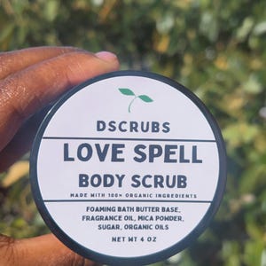 May include: A black jar with a white label that reads "DSCRUBS LOVE SPELL BODY SCRUB" and "MADE WITH 100% ORGANIC INGREDIENTS". The label also lists the ingredients: "FOAMING BATH BUTTER BASE, FRAGRANCE OIL, MICA POWDER, SUGAR, ORGANIC OILS" and "NET WT 4 OZ".