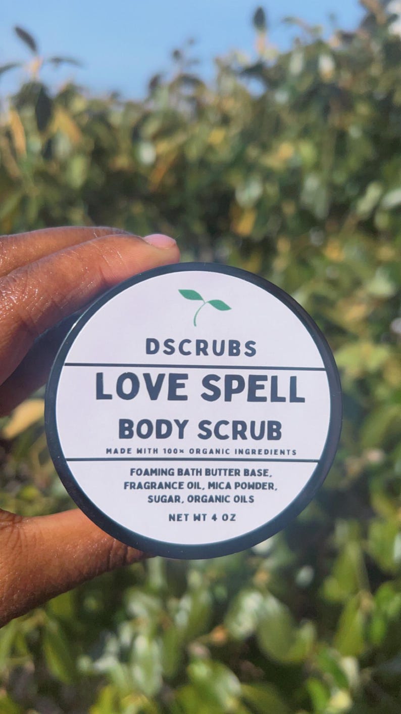 May include: A black jar with a white label that reads "DSCRUBS LOVE SPELL BODY SCRUB" and "MADE WITH 100% ORGANIC INGREDIENTS". The label also lists the ingredients: "FOAMING BATH BUTTER BASE, FRAGRANCE OIL, MICA POWDER, SUGAR, ORGANIC OILS" and "NET WT 4 OZ".