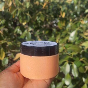 Op de afbeelding: Een lichaams scrub met perzik en bloesem geur in een doorzichtige pot met een zwarte deksel. De pot is gelabeld "Peach Blossom Body Scrub".