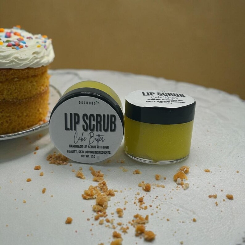 Puede incluir: Dos peque&ntilde;os frascos de exfoliante labial etiquetados como "Lip Scrub Cake Batter" con un producto amarillo en su interior. Los frascos est&aacute;n sobre una superficie blanca con algunas migajas esparcidas a su alrededor.
