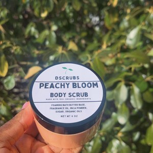 Op de afbeelding: Een witte pot met een zwarte deksel en een perzikkleurig etiket. Het etiket luidt "DSCRUBS PEACHY BLOOM BODY SCRUB MADE WITH 100% ORGANIC INGREDIENTS FOAMING BATH BUTTER BASE, FRAGRANCE OIL, MICA POWDER, SUGAR, ORGANIC OILS NET WT 4 OZ".