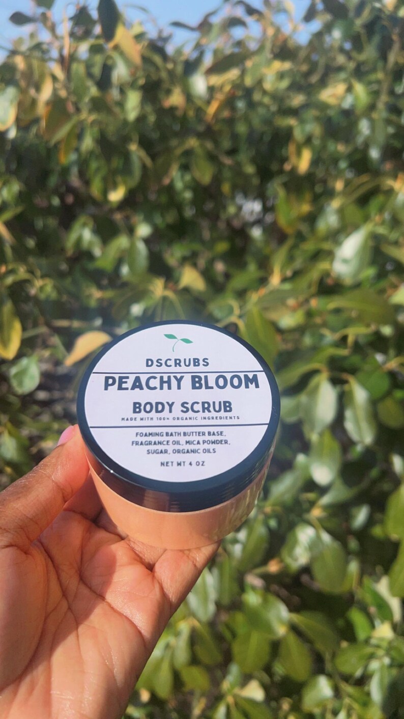 Op de afbeelding: Een witte pot met een zwarte deksel en een perzikkleurig etiket. Het etiket luidt "DSCRUBS PEACHY BLOOM BODY SCRUB MADE WITH 100% ORGANIC INGREDIENTS FOAMING BATH BUTTER BASE, FRAGRANCE OIL, MICA POWDER, SUGAR, ORGANIC OILS NET WT 4 OZ".