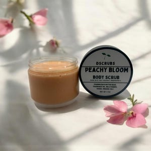 Op de afbeelding: Een perzikkleurige body scrub in een glazen pot met een zwarte deksel. De deksel heeft de tekst "DSCRUBS PEACHY BLOOM BODY SCRUB MADE WITH 100% ORGANIC INGREDIENTS FOAMING BATH BUTTER BASE, FRAGRANCE OIL, MICA POWDER, SUGAR, ORGANIC OILS NET WT 4 OZ".