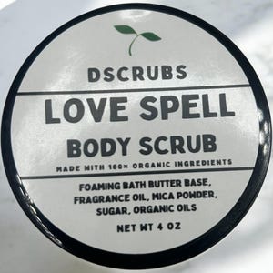 May include: A white jar with a black lid and a green plant logo. The jar is labeled "DSCRUBS LOVE SPELL BODY SCRUB MADE WITH 100% ORGANIC INGREDIENTS FOAMING BATH BUTTER BASE, FRAGRANCE OIL, MICA POWDER, SUGAR, ORGANIC OILS NET WT 4 OZ".