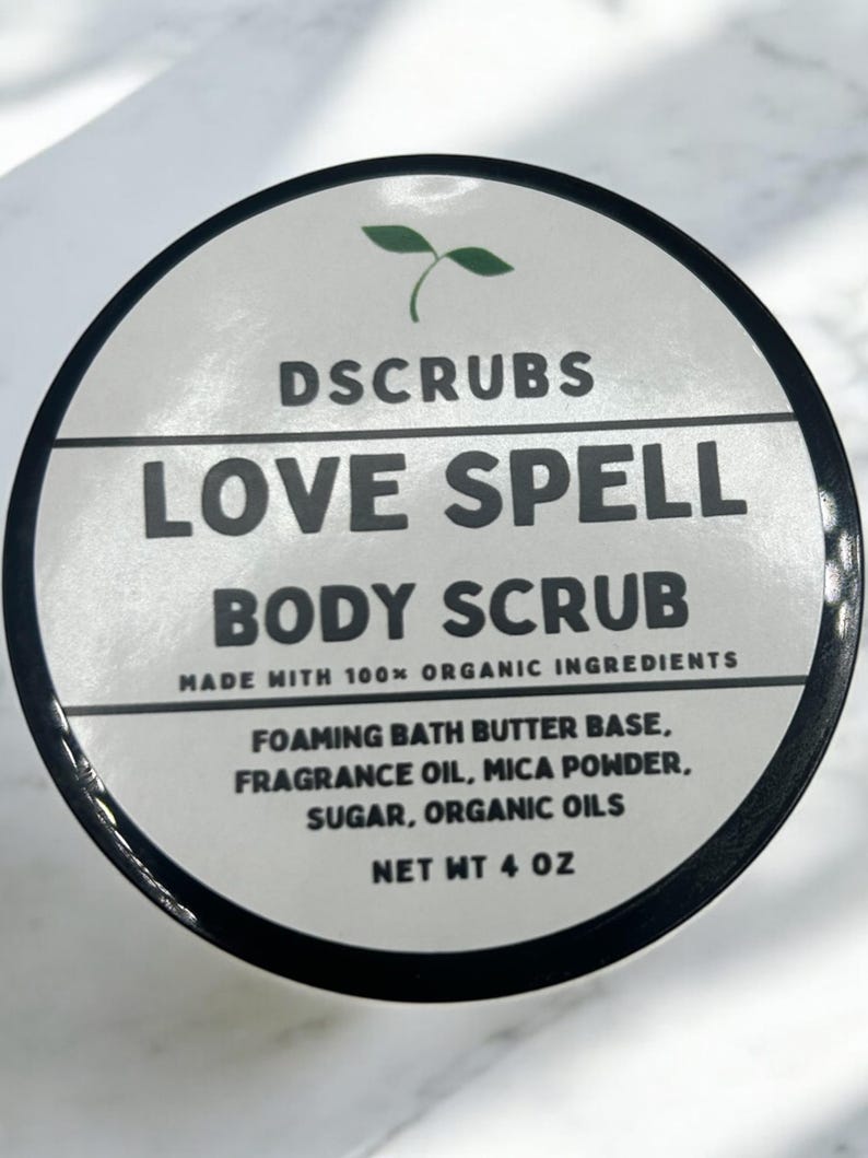 May include: A white jar with a black lid and a green plant logo. The jar is labeled "DSCRUBS LOVE SPELL BODY SCRUB MADE WITH 100% ORGANIC INGREDIENTS FOAMING BATH BUTTER BASE, FRAGRANCE OIL, MICA POWDER, SUGAR, ORGANIC OILS NET WT 4 OZ".
