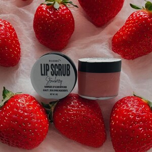 Puede incluir: Un peque&ntilde;o tarro de exfoliante labial de fresa con una etiqueta blanca que dice "USCRUBS LIP SCRUB Strawberry HANDMADE LIP SCRUB WITH HIGH QUALITY, SKIN LOVING INGREDIENTS. NET WT: 20Z". El tarro est&aacute; rodeado de fresas rojas.