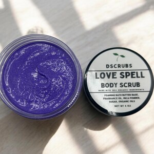 May include: A purple body scrub in a clear jar with a black lid. The jar is labeled "DSCRUBS LOVE SPELL BODY SCRUB MADE WITH 100% ORGANIC INGREDIENTS FOAMING BATH BUTTER BASE, FRAGRANCE OIL, MICA POWDER, SUGAR, ORGANIC OILS NET WT 4 OZ"