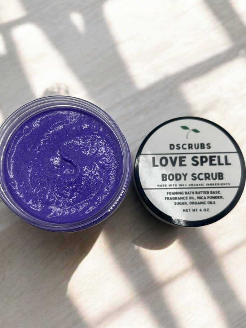 May include: A purple body scrub in a clear jar with a black lid. The jar is labeled "DSCRUBS LOVE SPELL BODY SCRUB MADE WITH 100% ORGANIC INGREDIENTS FOAMING BATH BUTTER BASE, FRAGRANCE OIL, MICA POWDER, SUGAR, ORGANIC OILS NET WT 4 OZ"