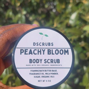 Op de afbeelding: Een zwarte pot met een wit etiket waarop staat "DSCRUBS PEACHY BLOOM BODY SCRUB MADE WITH 100% ORGANIC INGREDIENTS FOAMING BATH BUTTER BASE, FRAGRANCE OIL, MICA POWDER, SUGAR, ORGANIC OILS NET WT 4 OZ".
