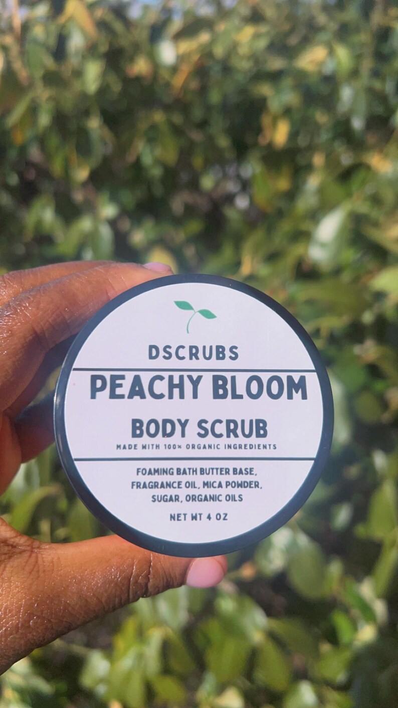 Op de afbeelding: Een zwarte pot met een wit etiket waarop staat "DSCRUBS PEACHY BLOOM BODY SCRUB MADE WITH 100% ORGANIC INGREDIENTS FOAMING BATH BUTTER BASE, FRAGRANCE OIL, MICA POWDER, SUGAR, ORGANIC OILS NET WT 4 OZ".