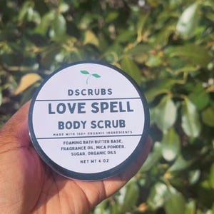 May include: A white jar with a black lid and a green leaf logo. The jar contains a body scrub called "Love Spell Body Scrub" made with 100% organic ingredients. The ingredients include foaming bath butter base, fragrance oil, mica powder, sugar, and organic oils. The jar is 4 oz.
