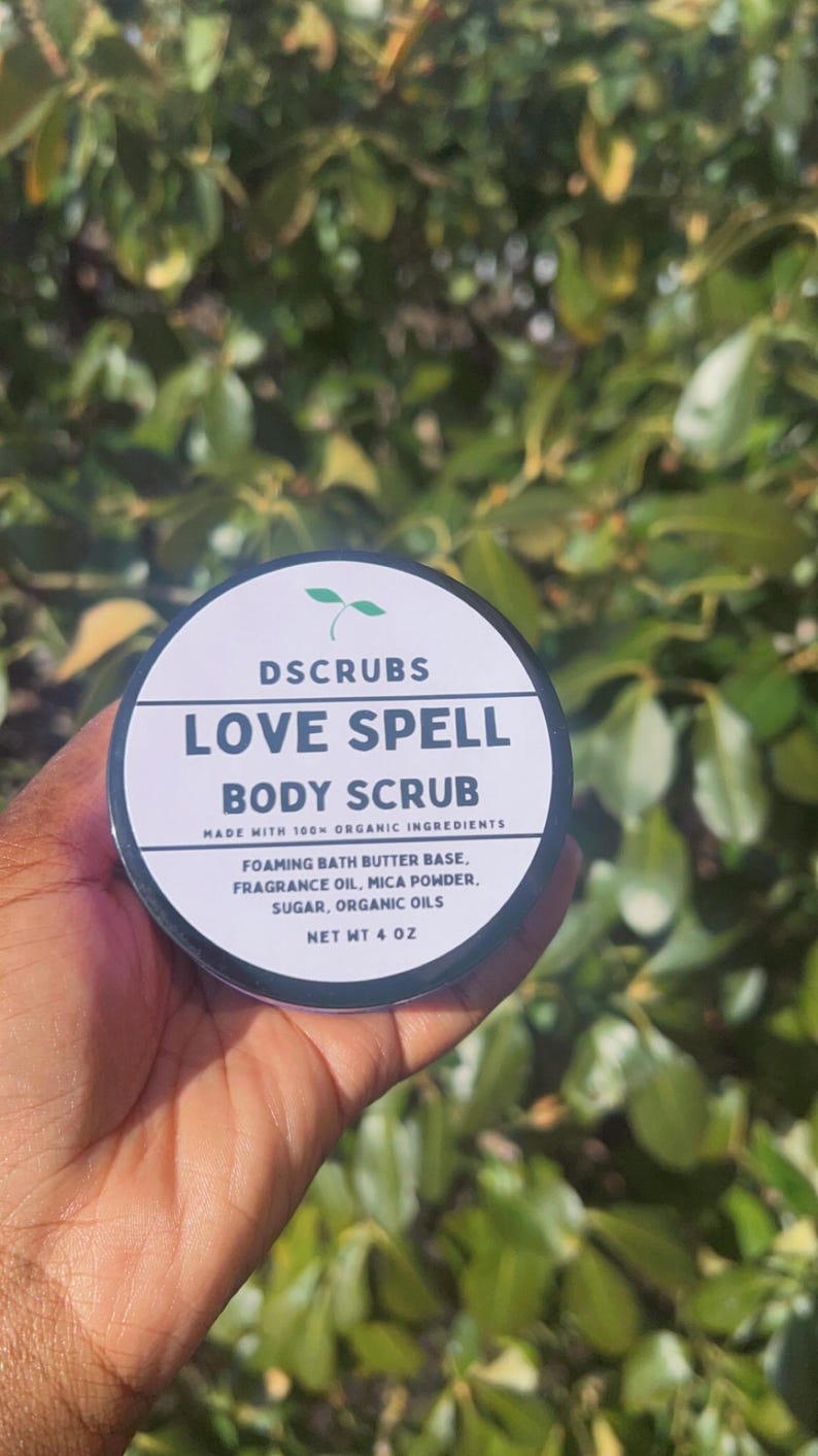 May include: A white jar with a black lid and a green leaf logo. The jar contains a body scrub called "Love Spell Body Scrub" made with 100% organic ingredients. The ingredients include foaming bath butter base, fragrance oil, mica powder, sugar, and organic oils. The jar is 4 oz.