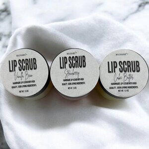Puede incluir: Tres peque&ntilde;os frascos de exfoliante labial con etiquetas blancas y texto negro. Las etiquetas dicen "Lip Scrub" con los sabores "Vanilla Bean", "Strawberry" y "Cake Batter".