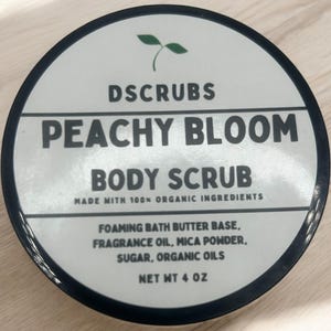 Op de afbeelding: Een witte pot met een zwarte deksel en een groen plantenlogo. De pot bevat een lichaams scrub met perzikgeur. Het etiket luidt "DSCRUBS PEACHY BLOOM BODY SCRUB MADE WITH 100% ORGANIC INGREDIENTS FOAMING BATH BUTTER BASE, FRAGRANCE OIL, MICA POWDER, SUGAR, ORGANIC OILS NET WT 4 OZ".