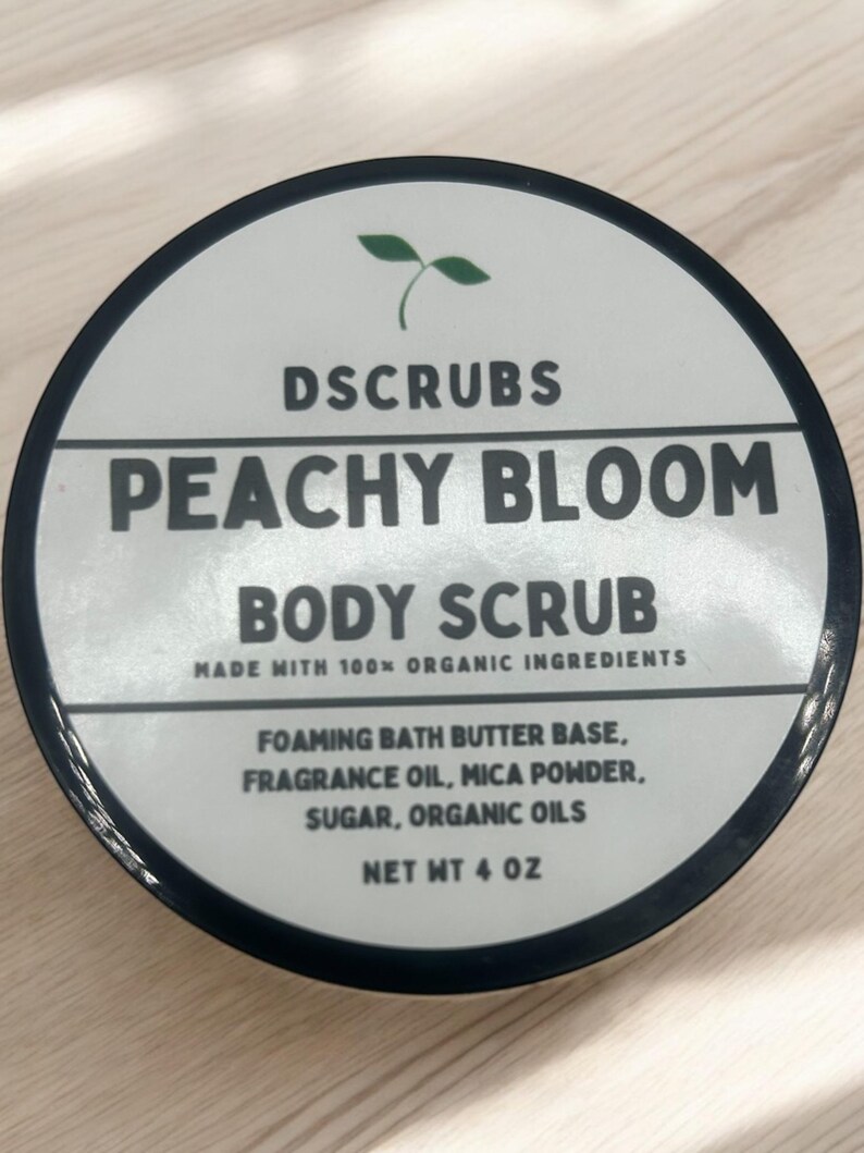 Op de afbeelding: Een witte pot met een zwarte deksel en een groen plantenlogo. De pot bevat een lichaams scrub met perzikgeur. Het etiket luidt "DSCRUBS PEACHY BLOOM BODY SCRUB MADE WITH 100% ORGANIC INGREDIENTS FOAMING BATH BUTTER BASE, FRAGRANCE OIL, MICA POWDER, SUGAR, ORGANIC OILS NET WT 4 OZ".