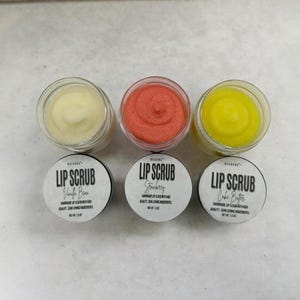 Puede incluir: Tres peque&ntilde;os frascos de vidrio con tapas blancas que contienen exfoliantes labiales. Los frascos est&aacute;n etiquetados con "Lip Scrub" con diferentes sabores: "Vanilla Bean", "Strawberry" y "Cake Batter".