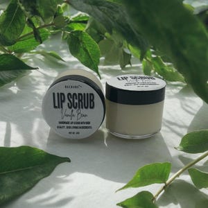 Puede incluir: Dos peque&ntilde;os frascos de exfoliante labial, uno etiquetado como "Lip Scrub Vanilla Bean" y el otro como "Lip Scrub". Ambos frascos son blancos con letras negras y est&aacute;n sobre una superficie blanca.