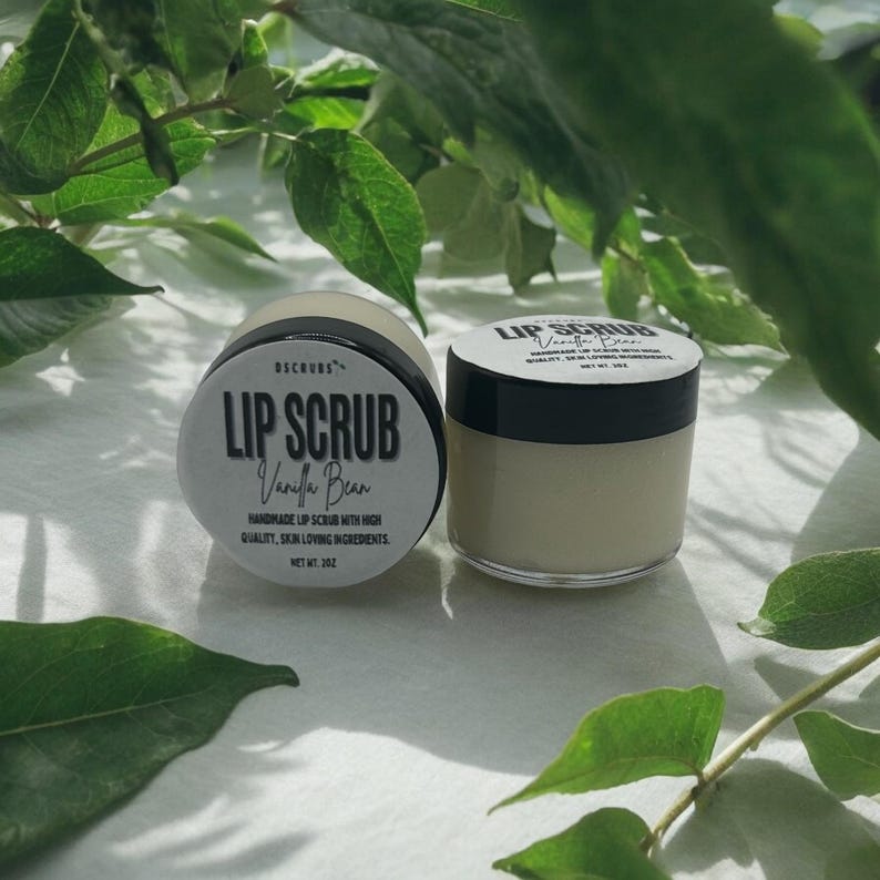Puede incluir: Dos peque&ntilde;os frascos de exfoliante labial, uno etiquetado como "Lip Scrub Vanilla Bean" y el otro como "Lip Scrub". Ambos frascos son blancos con letras negras y est&aacute;n sobre una superficie blanca.