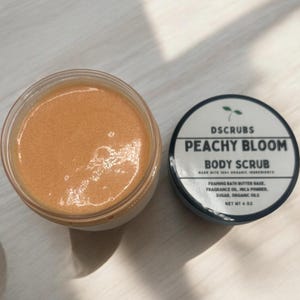 Op de afbeelding: Een perzikkleurige bodyscrub in een glazen pot met een zwarte deksel. De deksel heeft een wit etiket met de tekst "DSCRUBS PEACHY BLOOM BODY SCRUB MADE WITH 100% ORGANIC INGREDIENTS FOAMING BATH BUTTER BASE, FRAGRANCE OIL, MICA POWDER, SUGAR, ORGANIC OILS NET WT 4 OZ".