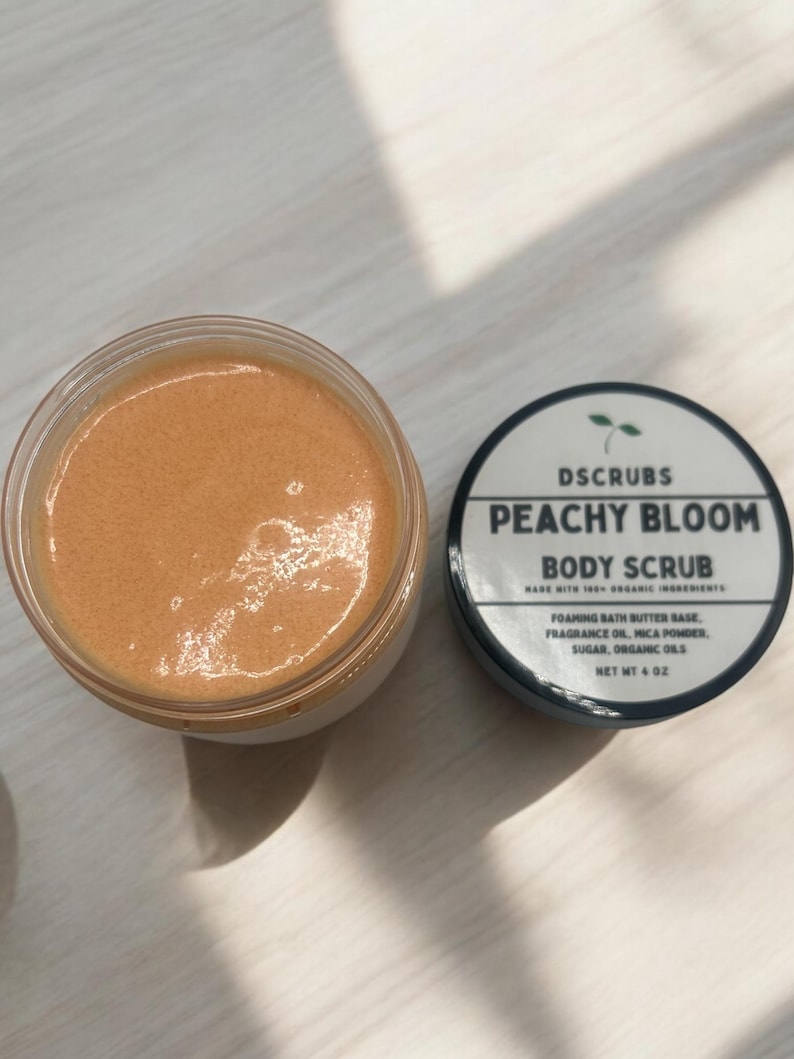 Op de afbeelding: Een perzikkleurige bodyscrub in een glazen pot met een zwarte deksel. De deksel heeft een wit etiket met de tekst "DSCRUBS PEACHY BLOOM BODY SCRUB MADE WITH 100% ORGANIC INGREDIENTS FOAMING BATH BUTTER BASE, FRAGRANCE OIL, MICA POWDER, SUGAR, ORGANIC OILS NET WT 4 OZ".