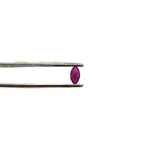 May include: A close-up of a small, marquise-shaped ruby gemstone held in tweezers. The ruby is a deep, rich red color.