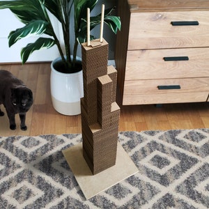 May include: A brown cardboard cat scratcher shaped like a skyscraper, with two wooden dowels at the top. A dark gray cat stands nearby on a patterned rug. The scratcher is on a light brown base.