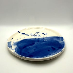 May include: A handmade ceramic plate with a white base and a bold blue painted design. The plate features a half-circle of blue paint and blue paint splatters. The plate is round with a slightly irregular edge.