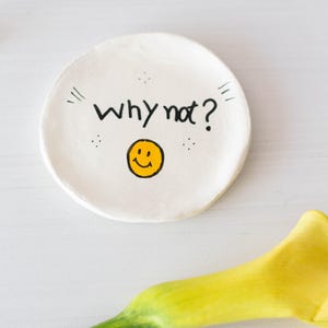 Puede incluir: Plato de cerámica blanco para anillos con el texto "Why not?" y una carita sonriente amarilla.