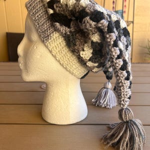 May include: A crocheted beanie hat featuring a variegated pattern of black, white, and gray yarn. The hat has a ribbed brim and a long, pointed tail that hangs down, finished with a large, multi-colored yarn tassel at the end of the point.
