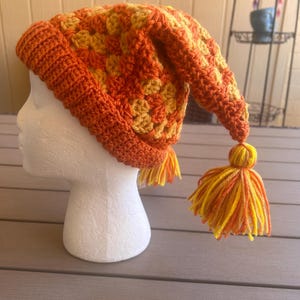 May include: A crocheted beanie hat featuring a vibrant orange and yellow granny square pattern. The hat has a folded ribbed brim in solid orange, a pointed top, and two dangling yarn tassels in matching orange and yellow hues.