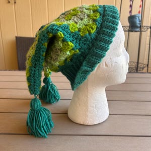 May include: A crocheted beanie hat featuring a granny square pattern in shades of green, lime, and teal. The hat has a long, pointed tail that hangs down the side, finished with two teal yarn tassels attached to the end of the point.