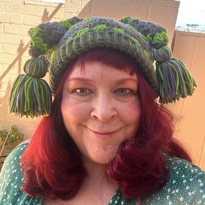 May include: A crocheted beanie hat featuring a checkered pattern in shades of dark green, light green, and grey. The hat has a ribbed brim and two large, dangling yarn tassels on each side, constructed from strands of green and grey yarn.