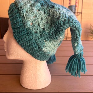 May include: A crocheted beanie in shades of teal, turquoise, and light blue, featuring a ribbed brim and a long, pointed tail that ends in a matching teal yarn tassel. The hat is constructed with a textured granny stitch pattern throughout.