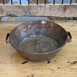 May include: A large copper bowl with two handles sits on a wooden surface. The bowl is slightly dented and has a patina.